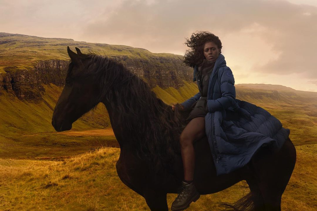 Woman in blue coat riding a black horse across a golden grassy field with rolling green hills under a sky with warm lighting.