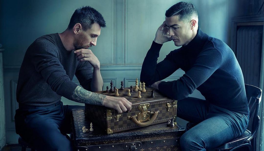 Lionel Messi and Cristiano Ronaldo face each other over a chess board placed on a Louis Vuitton trunk in a blue-toned indoor scene.