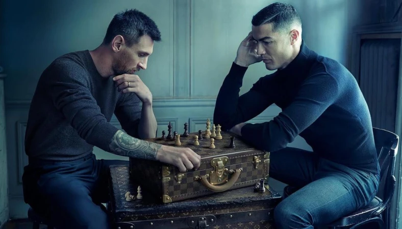 Lionel Messi and Cristiano Ronaldo face each other over a chess board placed on a Louis Vuitton trunk in a blue-toned indoor scene.