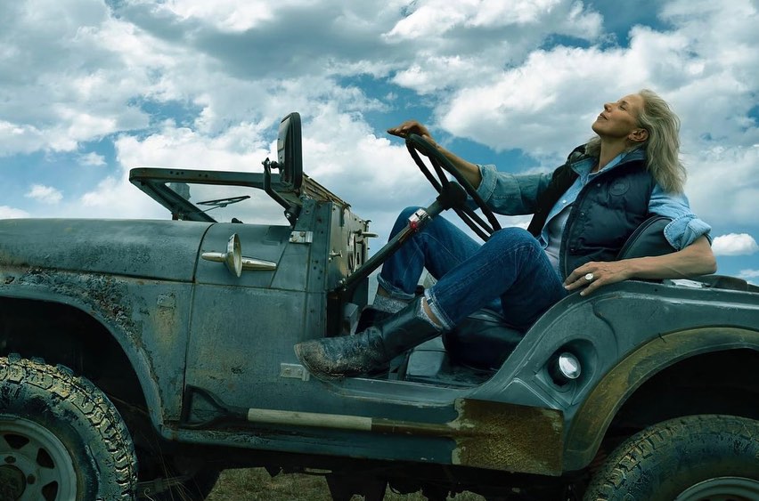 An older woman with grey hair sits in a weathered grey vintage jeep wearing blue jeans and a dark vest under a light blue shirt
