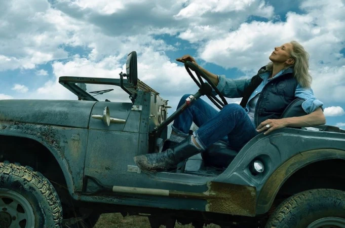 An older woman with grey hair sits in a weathered grey vintage jeep wearing blue jeans and a dark vest under a light blue shirt