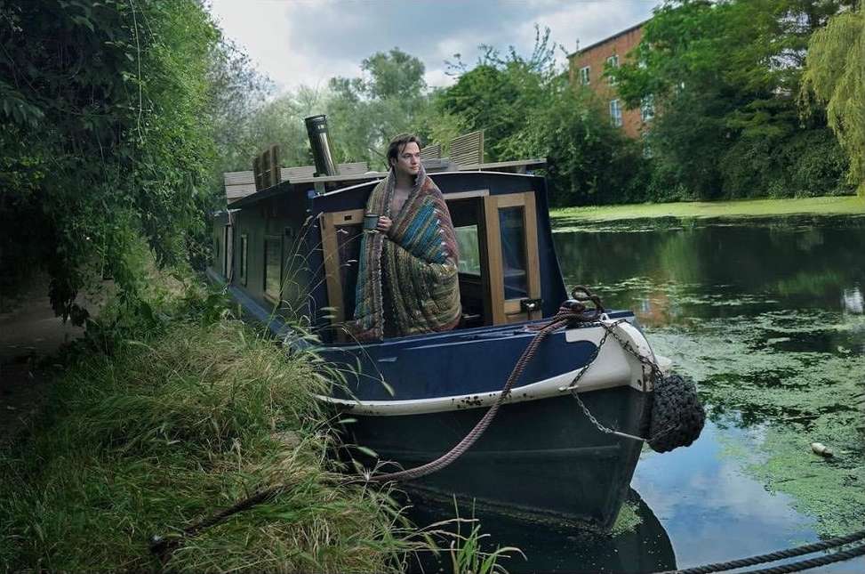 Adult male stands on a house boat wrapped in a colorful blanket, holding a mug, canal setting with green foliage and building in background.