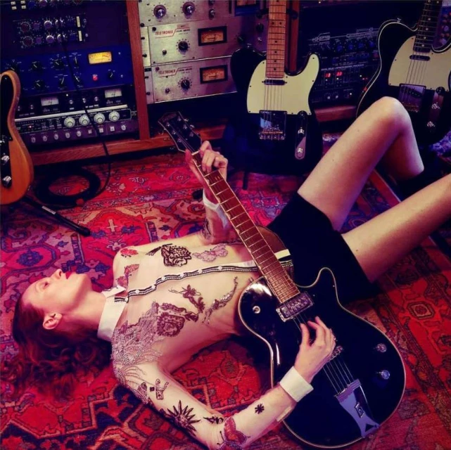 Young woman lying on an ornate rug in a music studio, holding a black electric guitar, surrounded by other guitars and sound equipment.
