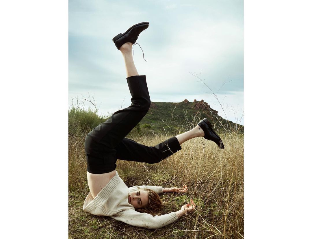 Woman in cream sweater and black pants does a headstand with feet pointing up in field with distant mountain in background.