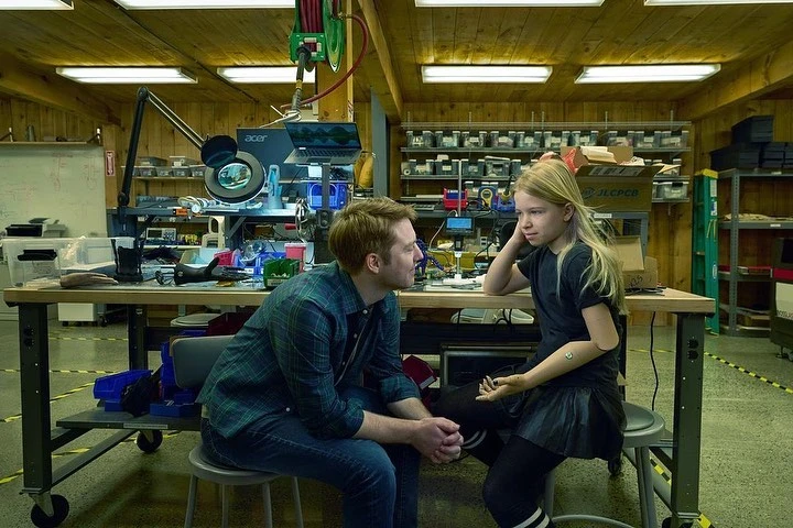 Young girl with prosthetic arm converses with adult male in workshop. The scene features fabrication equipment and wooden background.
