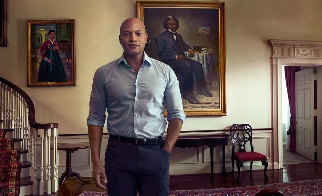 Bald man in a light blue shirt stands in a grand hallway with paintings and a staircase. The interior has an open doorway.