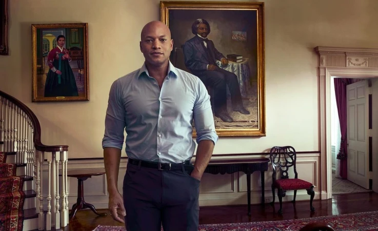 Portrait of a bald Black man standing in a traditional interior room with framed paintings on the wall behind him.