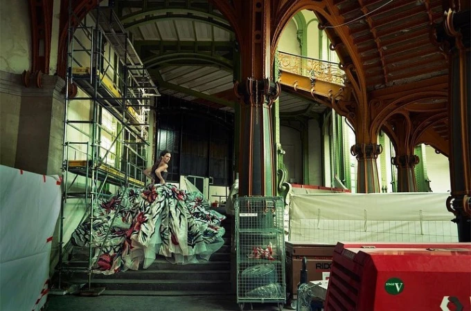Fashion photo shoot of woman in floral gown on stairs in unfinished architecture building with scaffolding and construction materials visible.