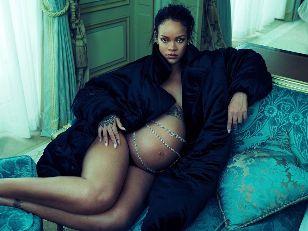 Pregnant Rihanna poses on a turquoise vintage couch wearing a black coat and a decorative silver belly chain with draped curtains.