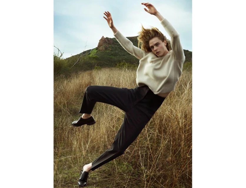 Fashion shot of a woman in a cream sweater and black pants mid-air in a grassy field with distant mountain. Editorial or lifestyle.