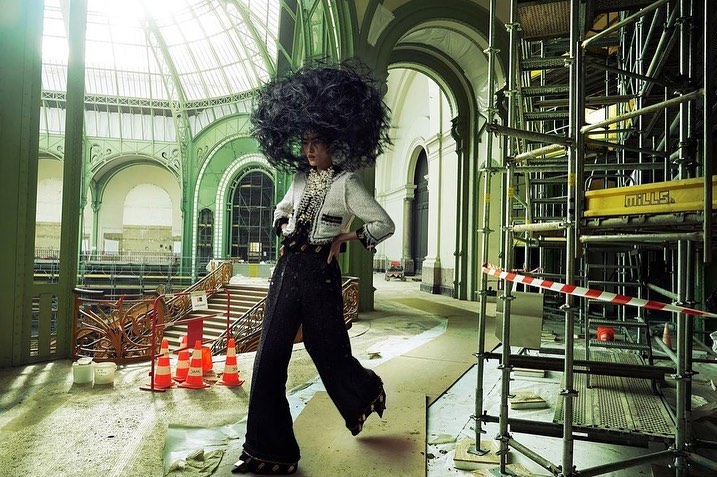 Fashionable woman in a large black headpiece poses in an ornate, under-construction hall. Architectural details & scaffolding surround her.