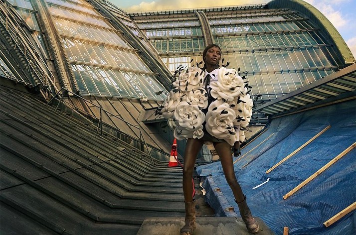 Fashionable African-American woman poses wearing a dress of white roses in an urban setting under a glass roof, showcasing style and design.
