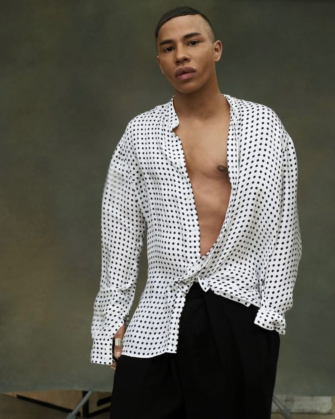 Man wearing a loose white polka dot button-down shirt and black trousers against a textured grey background.