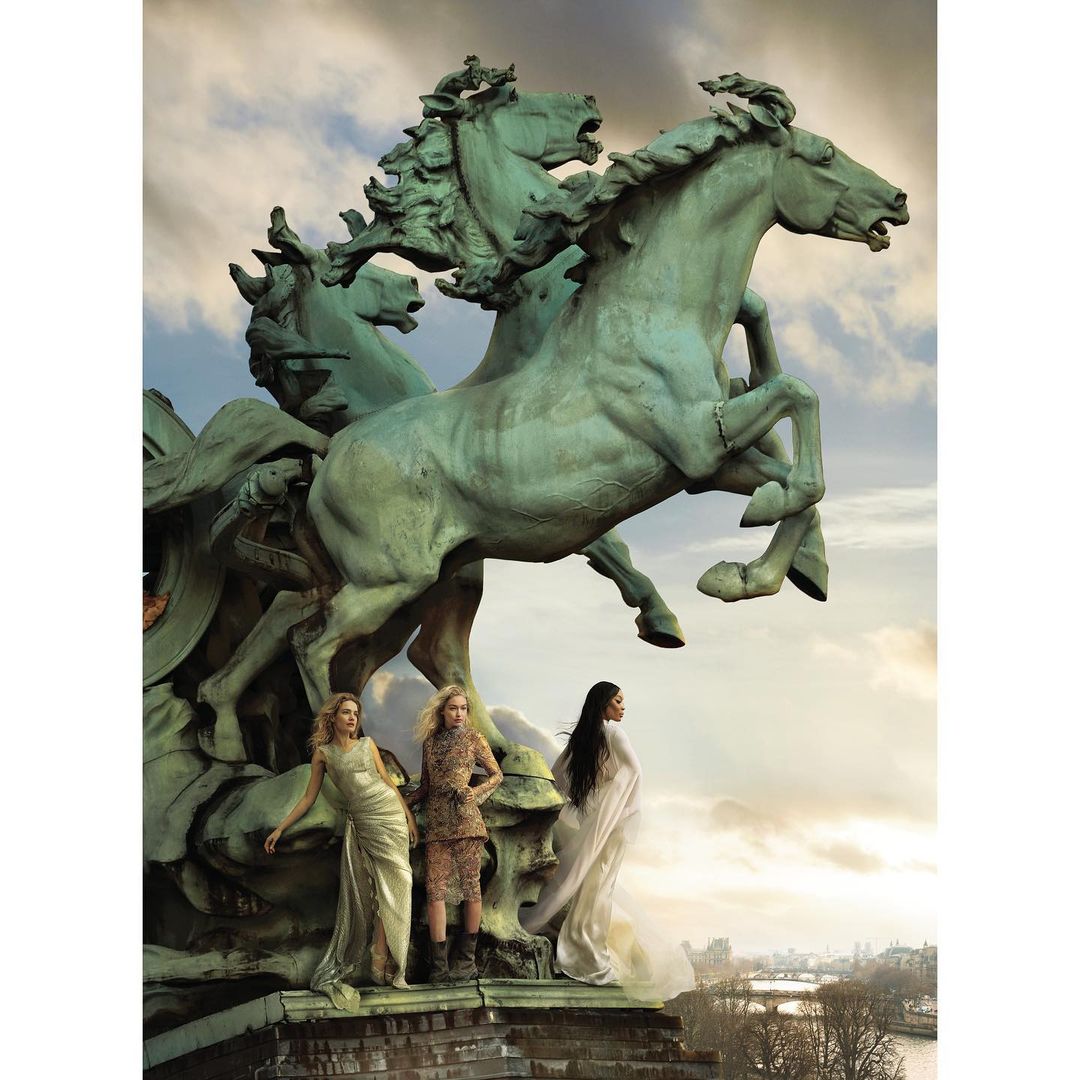 Three fashion models pose near a bronze sculpture of horses against a city and sky backdrop, showcasing editorial fashion and artistry.