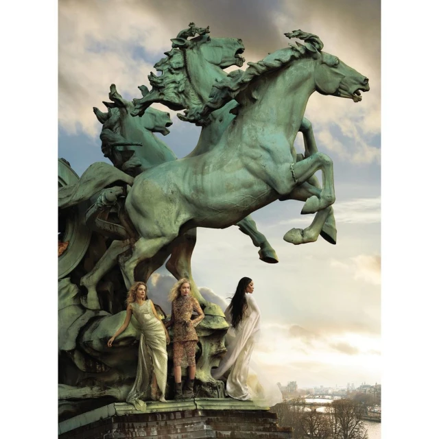 Three fashion models pose near a bronze sculpture of horses against a city and sky backdrop, showcasing editorial fashion and artistry.