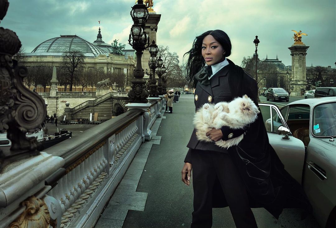 Fashion editorial shot of Naomi Campbell in Paris, holding a white cat, with the Pont Alexandre III bridge and architecture in the background.