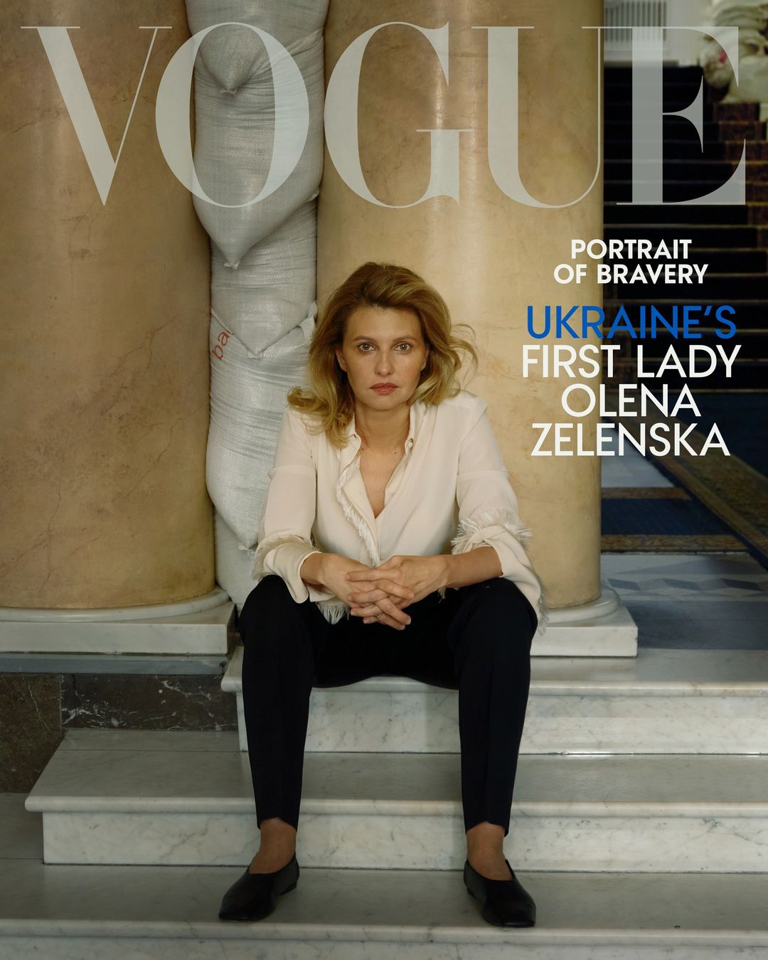 Olena Zelenska poses on marble steps for Vogue cover; portrait of bravery. She is wearing a white blouse and dark trousers, with column background.