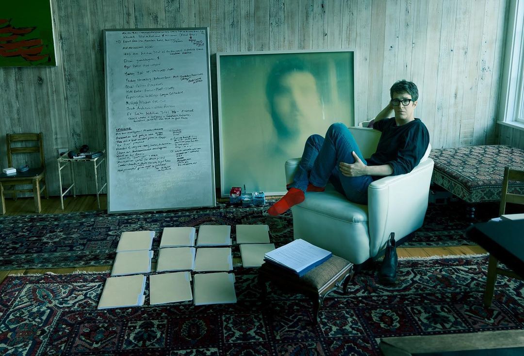 Adult male seated in an armchair in a room with a whiteboard, a framed image, and multiple stacks of paper folders on the floor.