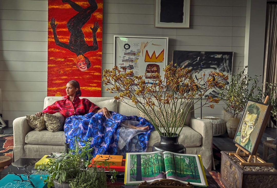 Elegant woman relaxes on a neutral sofa in a living room surrounded by modern art, colorful books, and decorative floral arrangements.