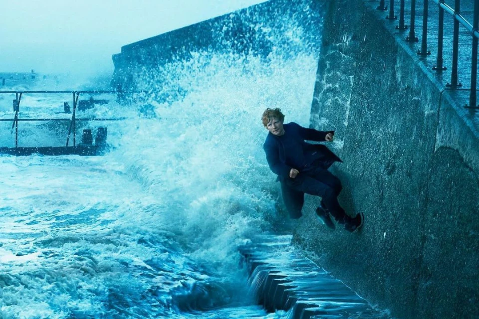 Adult male leans against a concrete sea wall with waves crashing and splashing against it on a cloudy day, wearing a navy overcoat.