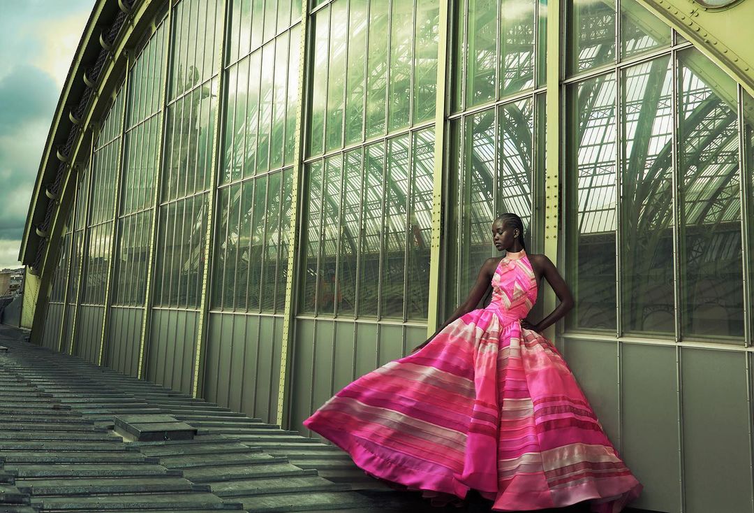 Fashion shot of a Black female model in a pink gown, posing in front of the facade of a greenhouse with a steel framework and glass panels.