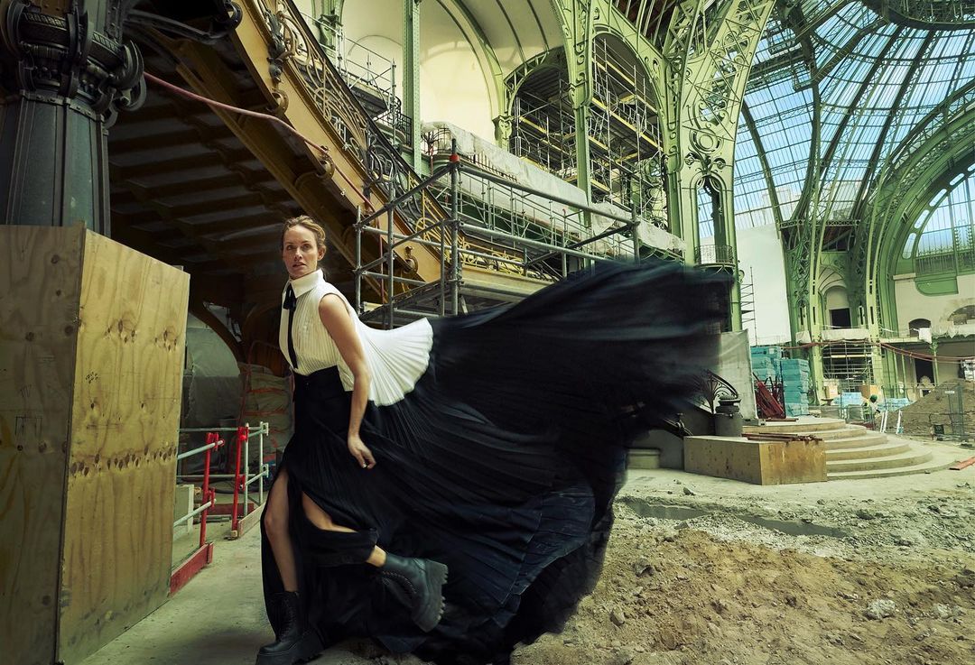 A woman in a black skirt and white shirt poses in a large hall with visible construction and scaffolding. The skirt is blowing in the wind.