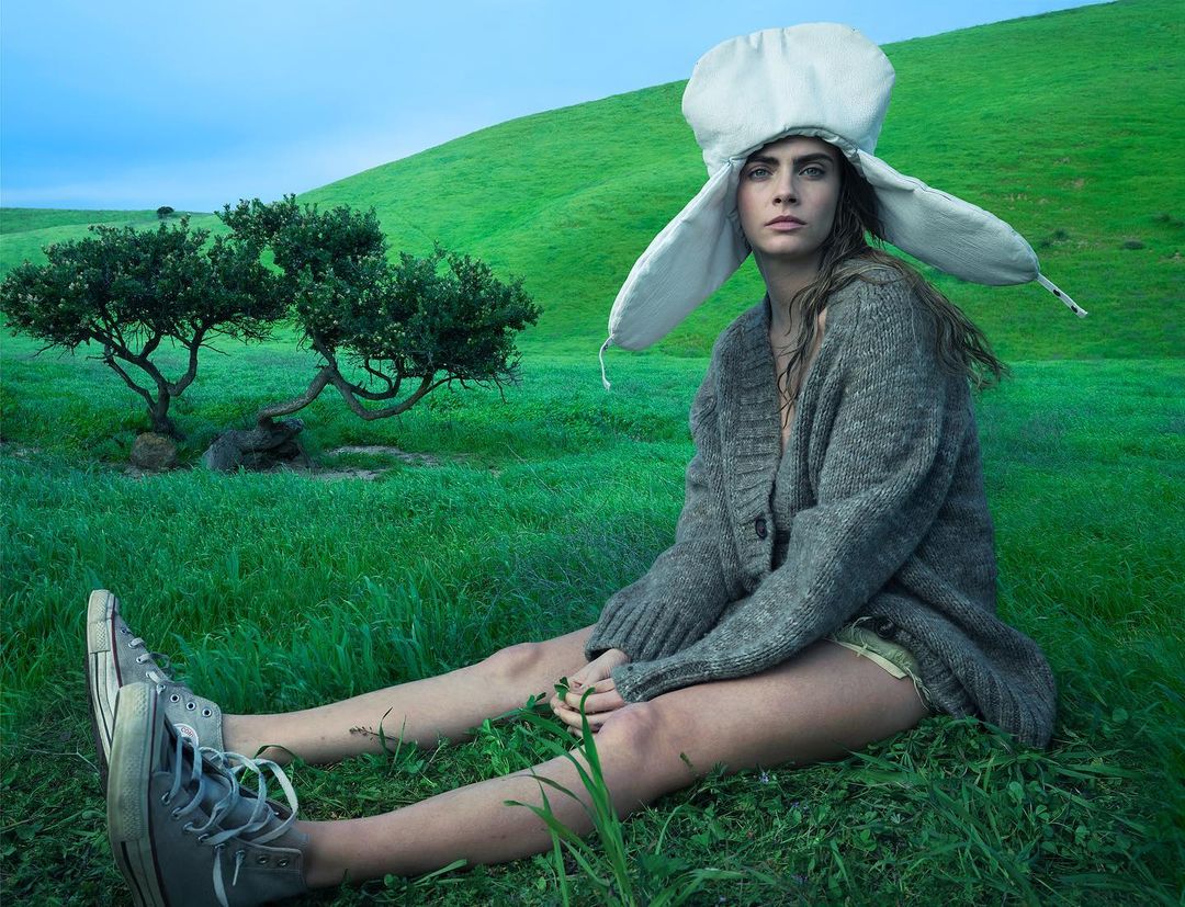 Woman with a whimsical hat sits in a green field. She wears a grey cardigan, and canvas shoes, creating a blend of nature and fashion.
