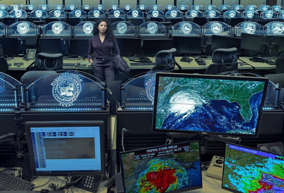 Woman stands in a control room with computer screens showing hurricane imagery, wearing a purple outfit and carrying a jacket.