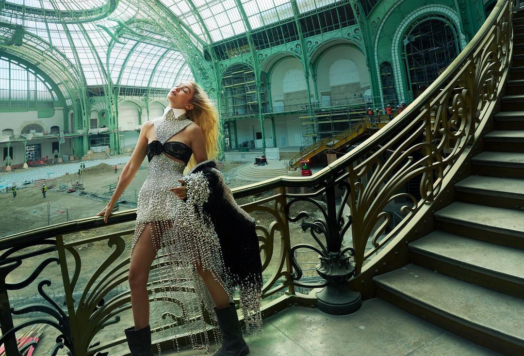 Fashion photo of a blonde model in a glittering dress standing on marble stairs inside a green and white building with metal scaffolding.