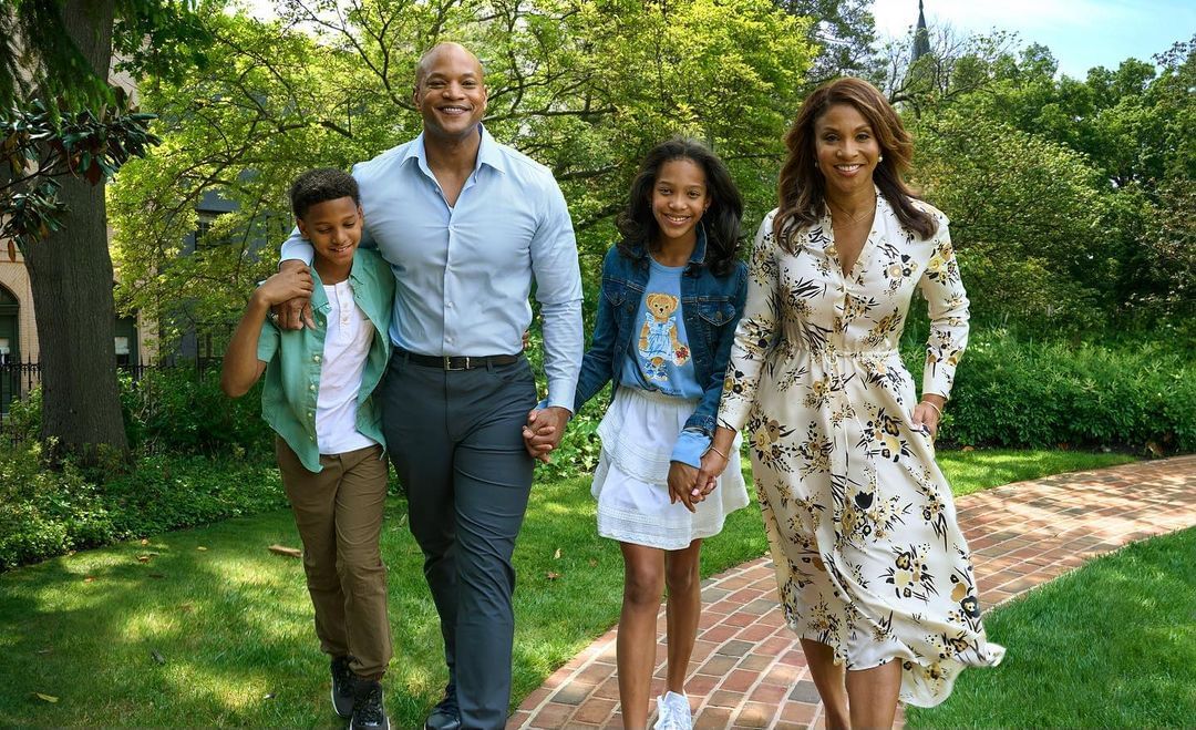 A family of four walks along a brick path in a green, lush park. The family consists of a mother, father, son, and daughter smiling.