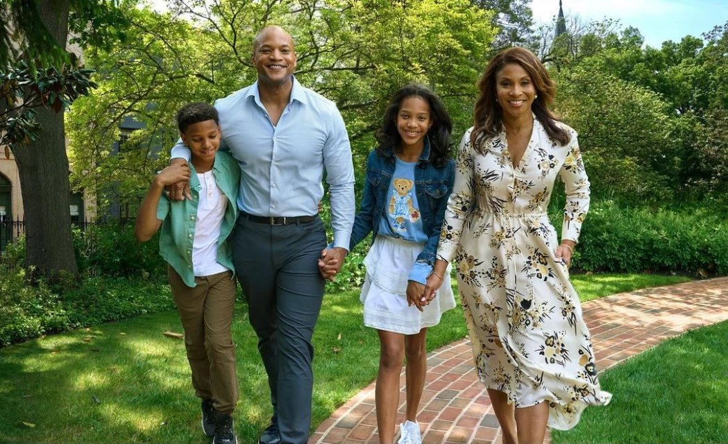 A family of four walks along a brick path in a green, lush park. The family consists of a mother, father, son, and daughter smiling.