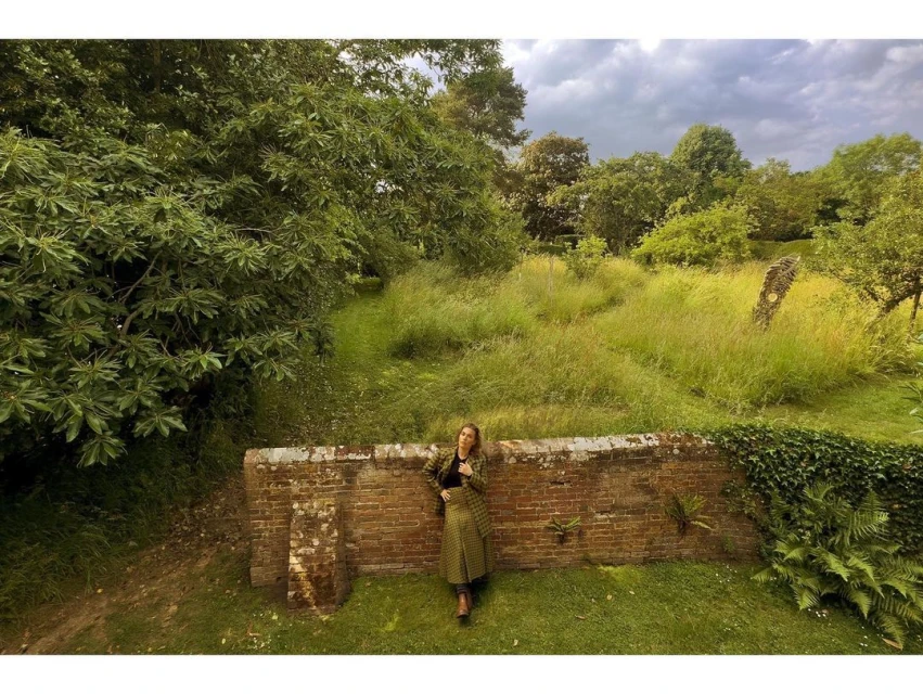 Adult female in a green patterned coat and black shoes standing on a grassy lawn in front of a brick wall with an overgrown field.