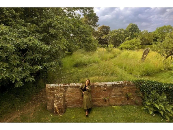Adult female in a green patterned coat and black shoes standing on a grassy lawn in front of a brick wall with an overgrown field.