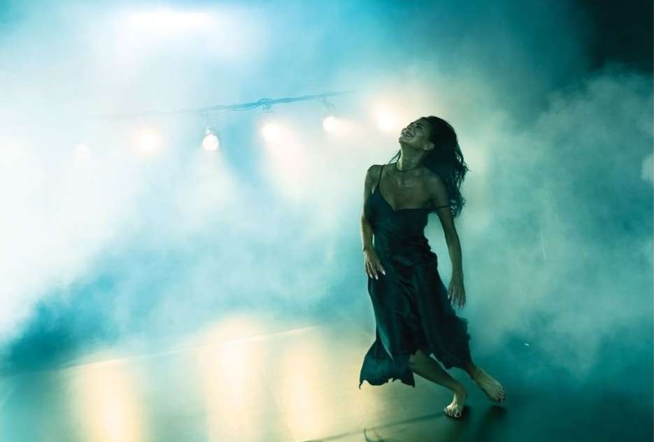 Woman in a black dress dancing in a smoky, atmospheric space with overhead stage lighting, showcasing dynamic movement and elegance.
