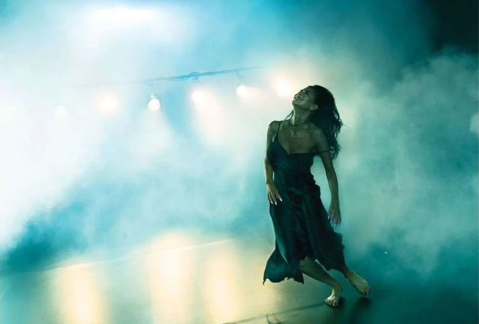 Woman in a black dress dancing in a smoky, atmospheric space with overhead stage lighting, showcasing dynamic movement and elegance.