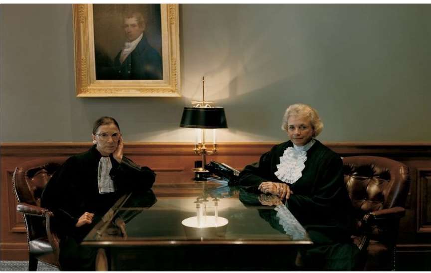 Two women seated at a table wearing black robes in an indoor environment. One woman rests her head on her hand.