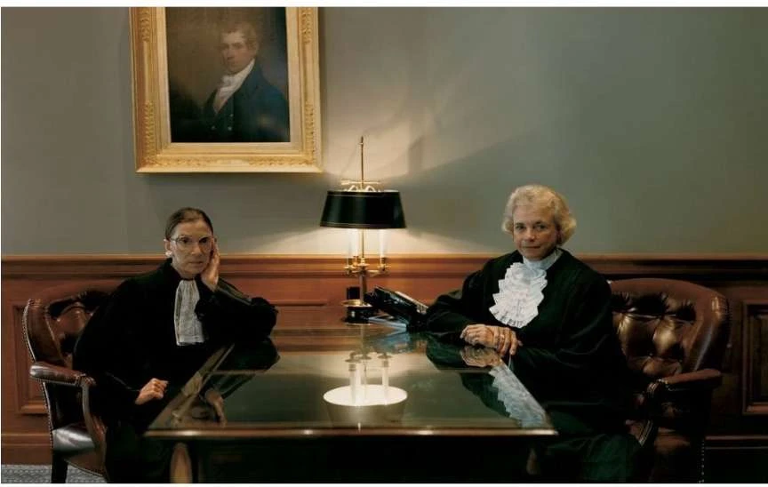 Two women seated at a table wearing black robes in an indoor environment. One woman rests her head on her hand.