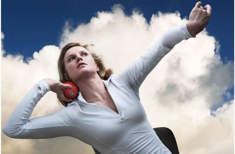 Female athlete prepares to throw a red shot put ball against a backdrop of a cloudy blue sky, wearing white athletic wear.