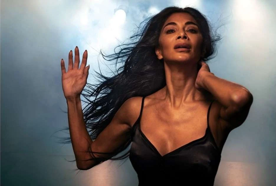 Portrait of an adult female in a black spaghetti-strap dress with long black hair and an outstretched hand in front of a diffused background.