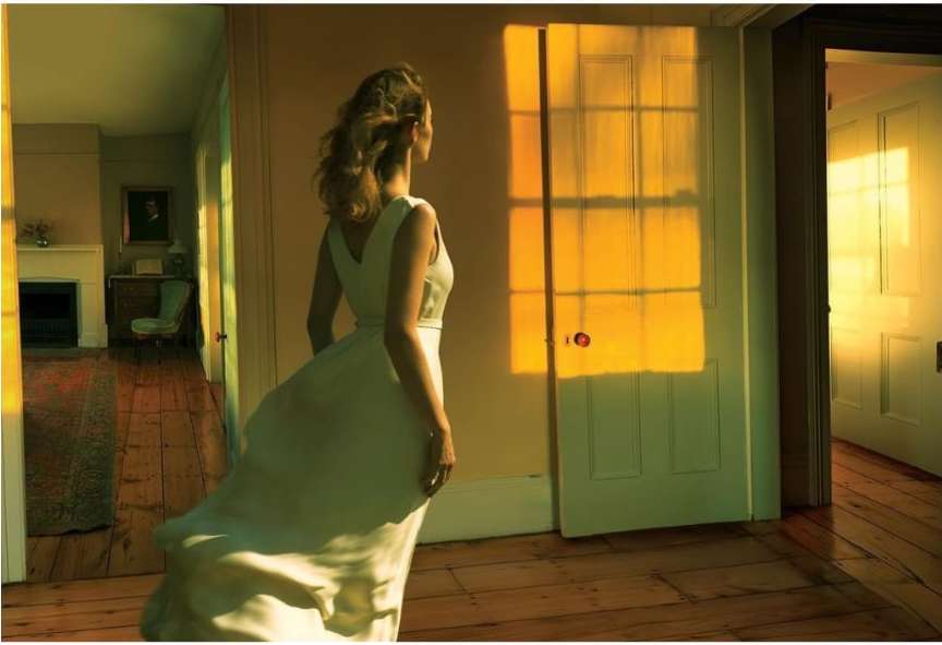 Woman in a white sleeveless dress standing in an interior hallway with warm golden sunlight streaming through an open door.