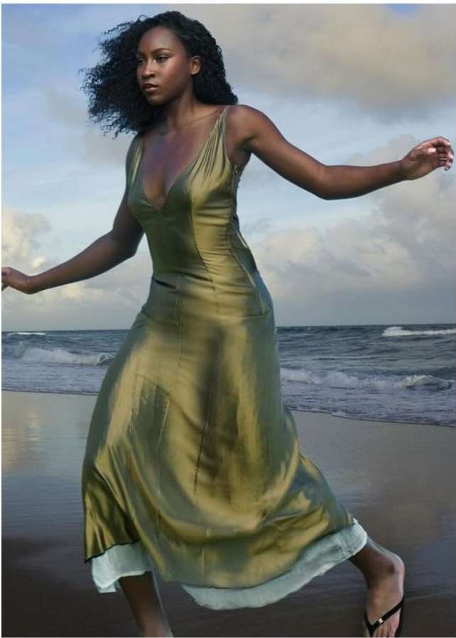 Black woman in a glossy olive green slip dress walking along a sandy beach under an overcast sky.