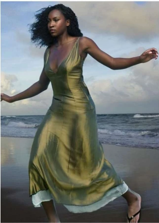Black woman in a glossy olive green slip dress walking along a sandy beach under an overcast sky.