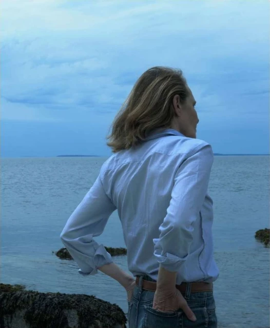 Adult woman stands on the rocky shore of a tranquil bay, gazing out at the calm water under a cloudy blue sky in a wide outdoor shot.