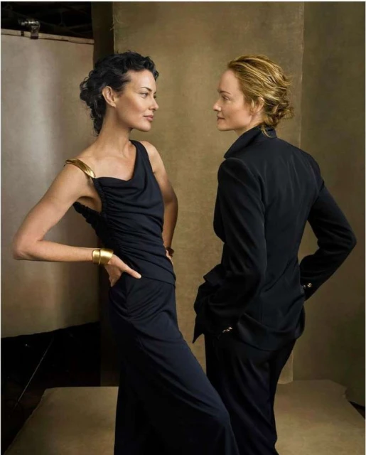 Two women stand indoors: one in a black gown with gold accents and one in a black suit, looking at each other in a studio setting.