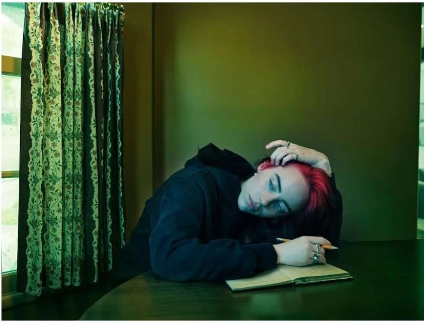 Person with red hair rests head on desk with notebook, wearing dark hoodie, with green wall and patterned curtains indoors.