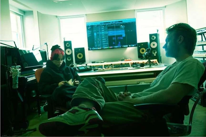 Two musicians in a music studio: One man sits in profile; another, with pink-streaked hair, reads in front of a soundboard and computer screen.