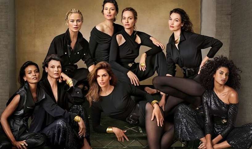 Diverse group of eight adult women in black clothing pose in a studio. The women are in a close huddle with neutral expressions.