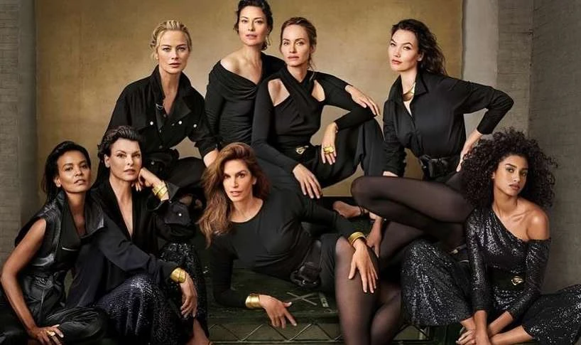 Diverse group of eight adult women in black clothing pose in a studio. The women are in a close huddle with neutral expressions.