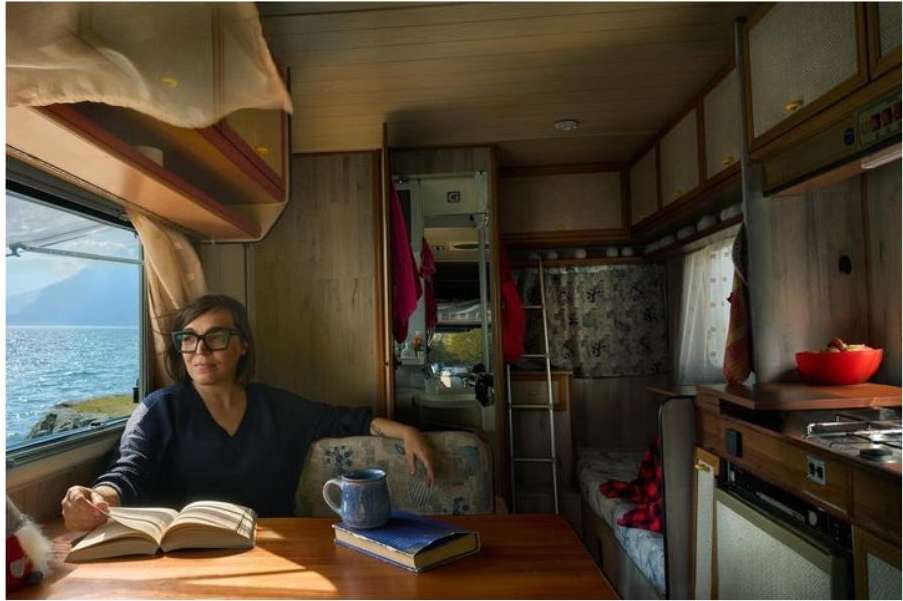 Woman in glasses reads a book at a table inside a camper by the window, with a view of the sea and mountains on a sunny day.