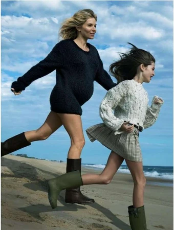 A pregnant woman and a child run barefoot on a sandy beach with blue skies. Both wear knit sweaters and tall boots, exuding carefree joy.